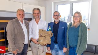 Vier Menschen stehen eng beieinander in einem hellen Raum mit einem hellen Fenster und lächeln für ein Foto. Ein Mann in einem weißen Hemd hält eine braune Tasche. Hinter ihnen zeigt eine Wand und ein Glasfenster einen Blick auf Bäume.