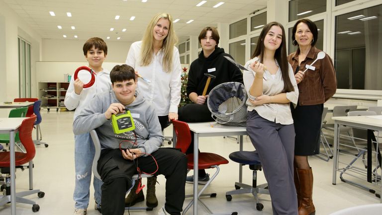 Eine Gruppe von Schülern und ein Lehrer posieren für ein Foto in einem Klassenzimmer mit verschiedenen Werkzeugen und Materialien auf einem Tisch.