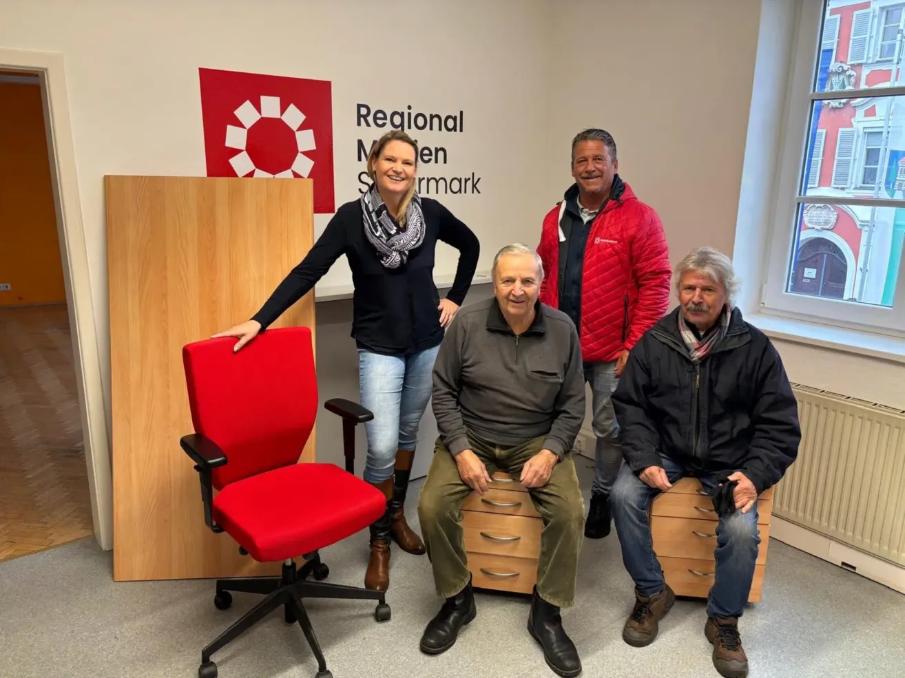 Fünf Personen posieren für ein Foto in einem Büro. Drei Männer sitzen auf Schubladen, während ein Mann steht. Eine Frau lehnt sich an einen Stuhl. Hinter ihnen steht eine Wand mit dem Text 'Regional Marken'.