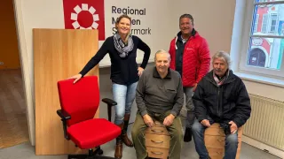 Fünf Personen posieren für ein Foto in einem Büro. Drei Männer sitzen auf Schubladen, während ein Mann steht. Eine Frau lehnt sich an einen Stuhl. Hinter ihnen steht eine Wand mit dem Text 'Regional Marken'.
