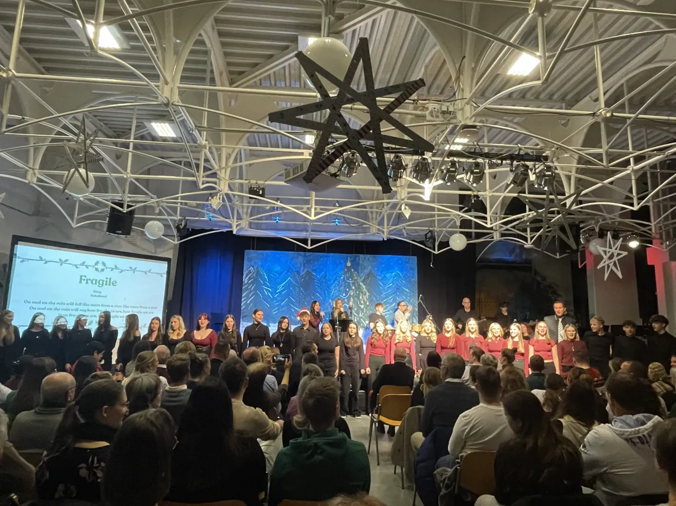 Ein Chor tritt auf einer Bühne in einem großen Raum auf, mit einer Projektionsleinwand hinter ihnen, die einen Weihnachtsbaum zeigt. Das Publikum sitzt in Stühlen vor der Bühne, und einige stehen.