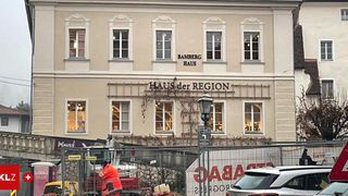 Ein Gebäude mit mehreren Fenstern und einem Schild mit der Aufschrift Bamberg Haus befindet sich im Bau. Ein Arbeiter in einer orangefarbenen Weste steht in der Nähe eines Fahrzeugs. Ein Schild mit rotem Text und dem Wort "SAUNA" ist sichtbar.