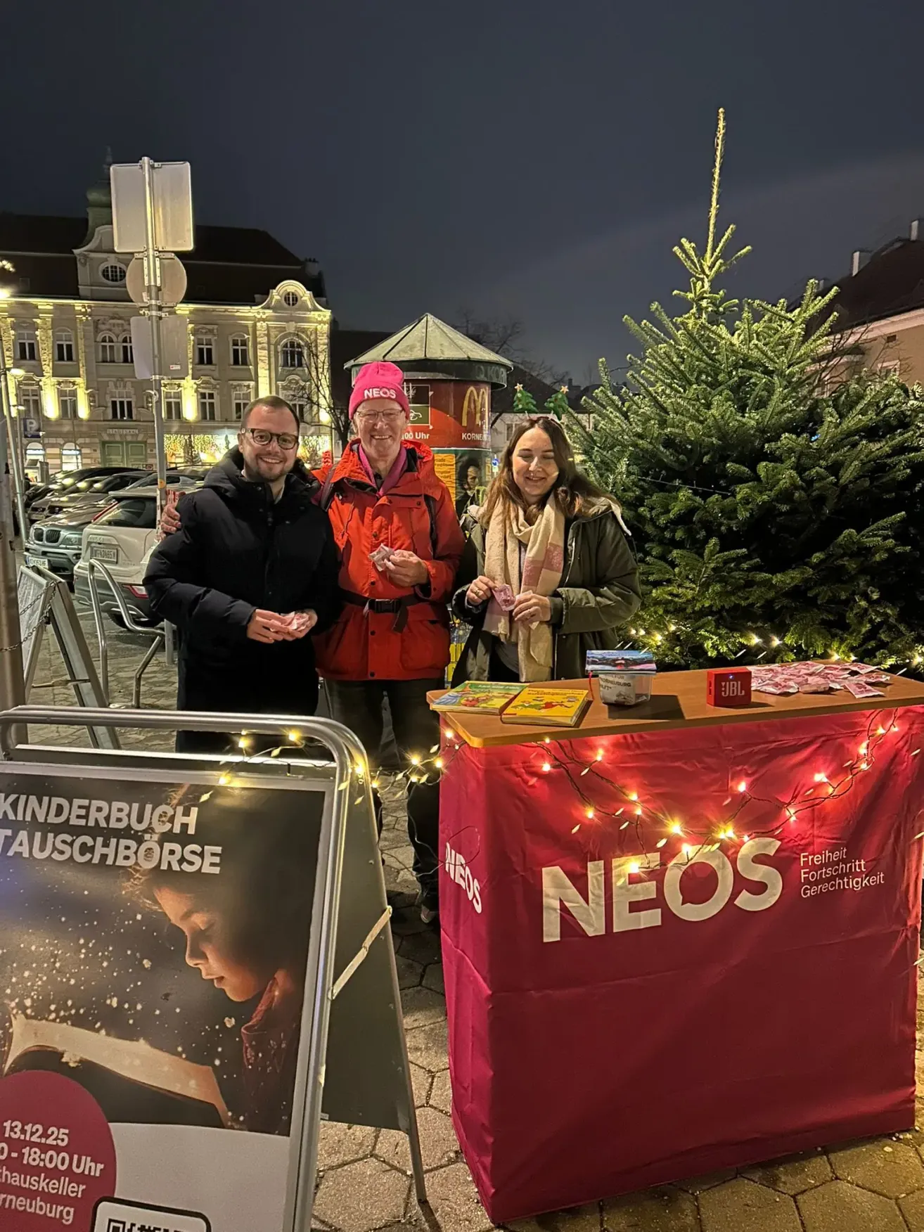 Drei Personen stehen neben einem roten Tisch mit dem Wort Neos darauf. Sie lächeln für die Kamera. Hinter ihnen ist ein McDonalds und ein großer Baum.