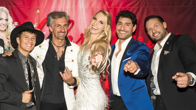 Vier lächelnde Personen posieren für ein Bild, darunter eine Frau in einem silbernen Kleid und Männer in Anzügen, einer in Blau und der andere in Weiß. Sie stehen vor einem rosa Hintergrund.