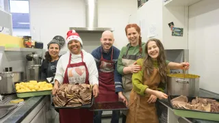 Fünf Personen in einer Küche mit Schürzen, eine Person hält eine Schale mit geschnittenem Fleisch. Hinter ihnen sind eine Uhr, ein Regal und ein Herd zu sehen.