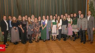 Eine Gruppe von Frauen und einem Mann posiert für ein Foto vor einem Vorhang, einige in traditioneller Kleidung, stehen auf einem Holzboden.