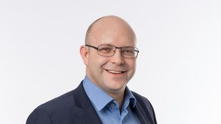 Ein kahlköpfiger Mann mit Brille lächelt und trägt ein blaues Hemd und eine dunkle Jacke vor weißem Hintergrund.