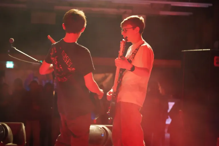 Zwei Jungen spielen Gitarren auf einer Bühne unter roter Beleuchtung. Der Junge links trägt ein schwarzes T-Shirt. Der Junge rechts trägt ein weißes T-Shirt und eine Brille. Vor ihnen stehen ein Lautsprecher und ein Mikrofonständer.