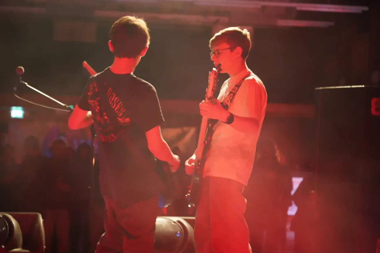 Zwei Jungen spielen Gitarren auf einer Bühne unter roter Beleuchtung. Der Junge links trägt ein schwarzes T-Shirt. Der Junge rechts trägt ein weißes T-Shirt und eine Brille. Vor ihnen stehen ein Lautsprecher und ein Mikrofonständer.