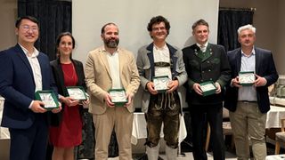 Eine Gruppe von Menschen in formeller Kleidung, die grüne Schachteln halten, stehen bei einer Veranstaltung zusammen. Sie lächeln und posieren für ein Foto. Hinter ihnen befindet sich eine weiße Wand mit schwarzen Vorhängen. Es gibt einen Tisch mit weißer Tischdecke.