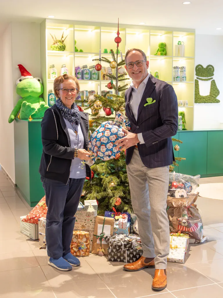 Ein Mann und eine Frau stehen neben einem Weihnachtsbaum mit Geschenken. Sie lächeln und halten ein Geschenk. Im Hintergrund befinden sich Regale mit Spielzeug und eine große Froschfigur.
