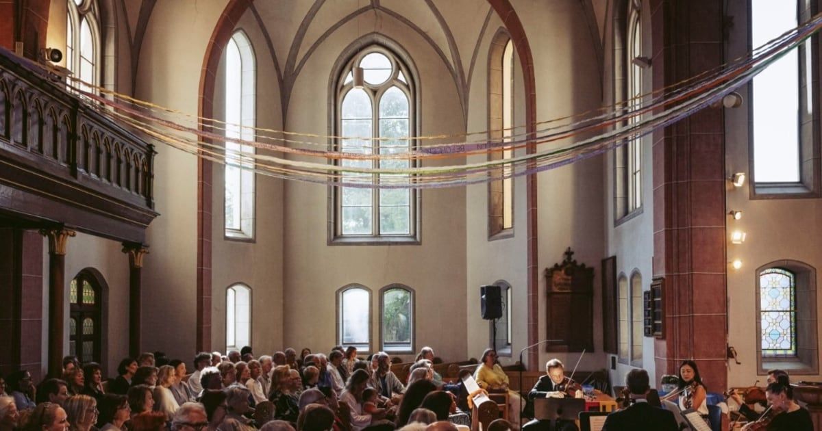 Ein Geiger spielt ein Solo in einer Kirche, vor einer versammelten Menschenmenge. Der Raum hat gewölbte Fenster und Dekorationen.