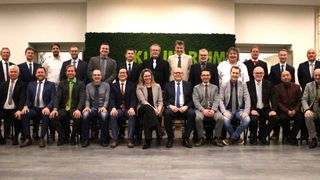 Eine Gruppe von Fachleuten in formeller Kleidung sitzt vor einem grünen Hintergrund. Sie scheinen für ein Foto zu posieren.