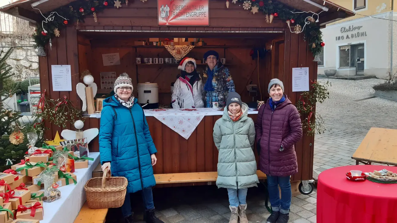 Fünf Frauen in Wintermänteln stehen vor einem festlichen Stand. Sie lächeln und scheinen für ein Foto zu posieren. Der Stand hat ein Schild mit der Aufschrift 'Obergailtaler Trachtengrufte'. Dahinter steht ein Regal mit Gegenständen, und eine Person sitzt drinnen.