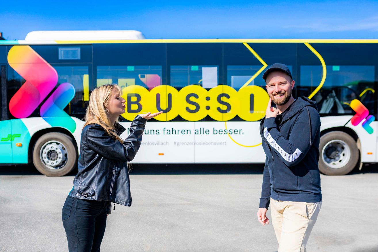 Ein Mann und eine Frau stehen vor einem Bus mit der Aufschrift Bus:SI. Sie scheinen für ein Foto zu posieren.