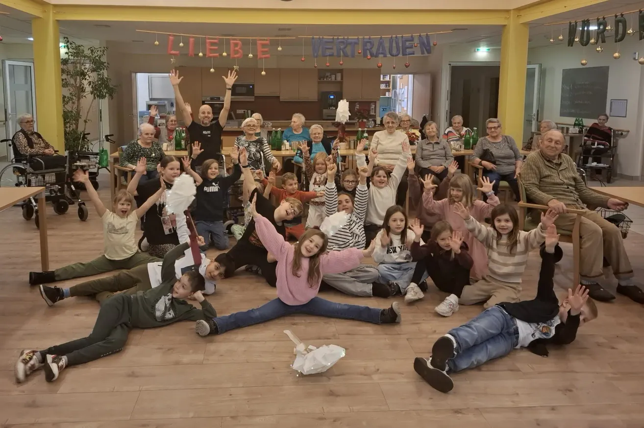 Eine Gruppe von Menschen versammelt sich in einem Raum, einige sitzen und andere stehen und feiern. Viele Kinder heben ihre Hände. Über ihnen hängt ein Banner mit den Worten 'Liebe Vertrauen'.