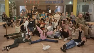 Eine Gruppe von Menschen versammelt sich in einem Raum, einige sitzen und andere stehen und feiern. Viele Kinder heben ihre Hände. Über ihnen hängt ein Banner mit den Worten 'Liebe Vertrauen'.