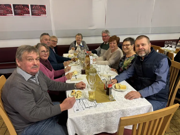 Eine Gruppe von Menschen sitzt um einen Tisch mit Essen, Getränken und Weingläsern. Sie scheinen eine Mahlzeit gemeinsam zu genießen.