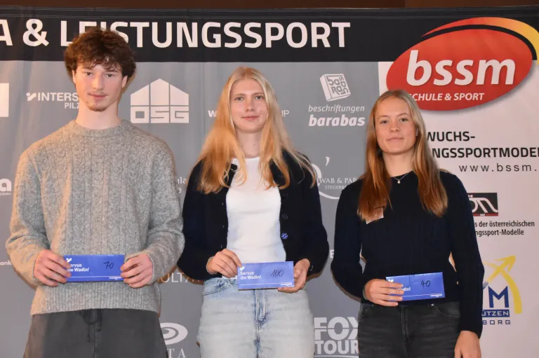 Drei junge Menschen stehen zusammen und halten blaue Urkunden. Sie befinden sich wahrscheinlich bei einer Veranstaltung von Stiftungssport. Die beiden Frauen tragen schwarze Oberteile und der Mann ein graues Sweatshirt. Im Hintergrund sind ein Logo und Text zu Stiftungssport zu sehen.