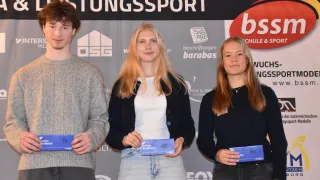 Drei junge Menschen stehen zusammen und halten blaue Urkunden. Sie befinden sich wahrscheinlich bei einer Veranstaltung von Stiftungssport. Die beiden Frauen tragen schwarze Oberteile und der Mann ein graues Sweatshirt. Im Hintergrund sind ein Logo und Text zu Stiftungssport zu sehen.