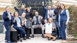 Eine Gruppe von Menschen posiert für ein Foto im Freien. Sie sitzen auf einer Bank und lächeln. Sie halten ein Zertifikat in den Händen. Dahinter befindet sich ein schwarzer Banner mit dem Text 'Herzliche Willkommen'. Sie tragen alle formelle Kleidung. Im Hintergrund befinden sich zwei Bäume.