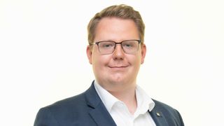 Ein Mann in einem dunkelblauen Anzug und weißem Hemd posiert für ein Bild vor weißem Hintergrund. Er trägt eine schwarze Brille und hat kurze, braune Haare.