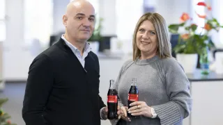 Ein Mann und eine Frau stehen in einem Büro, lächeln und halten Coca-Cola-Flaschen.
