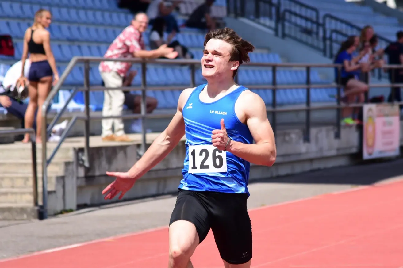 Ein männlicher Athlet läuft auf einer Laufbahn, die Nummer 126 auf seinem Trikot tragend, und zeigt den Daumen nach oben. Dahinter steht ein älterer Mann auf einer Treppe.