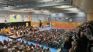 Ein großer Innenraum beherbergt ein klassisches Orchesterkonzert. Die Bühne ist mit Instrumenten ausgestattet, und das Publikum sitzt vor der Bühne auf Stühlen. Die Decke ist hoch mit Beleuchtungskörpern.