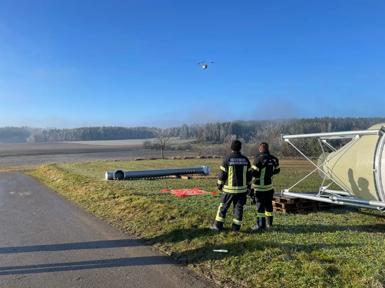 Zwei Feuerwehrleute stehen auf einem Grasfeld in der Nähe einer Straße. Ein Drohnen fliegt darüber. Eine große Rohr liegt auf dem Gras, und eine große Metallstruktur ist rechts. Bäume und ein Wald sind im Hintergrund.