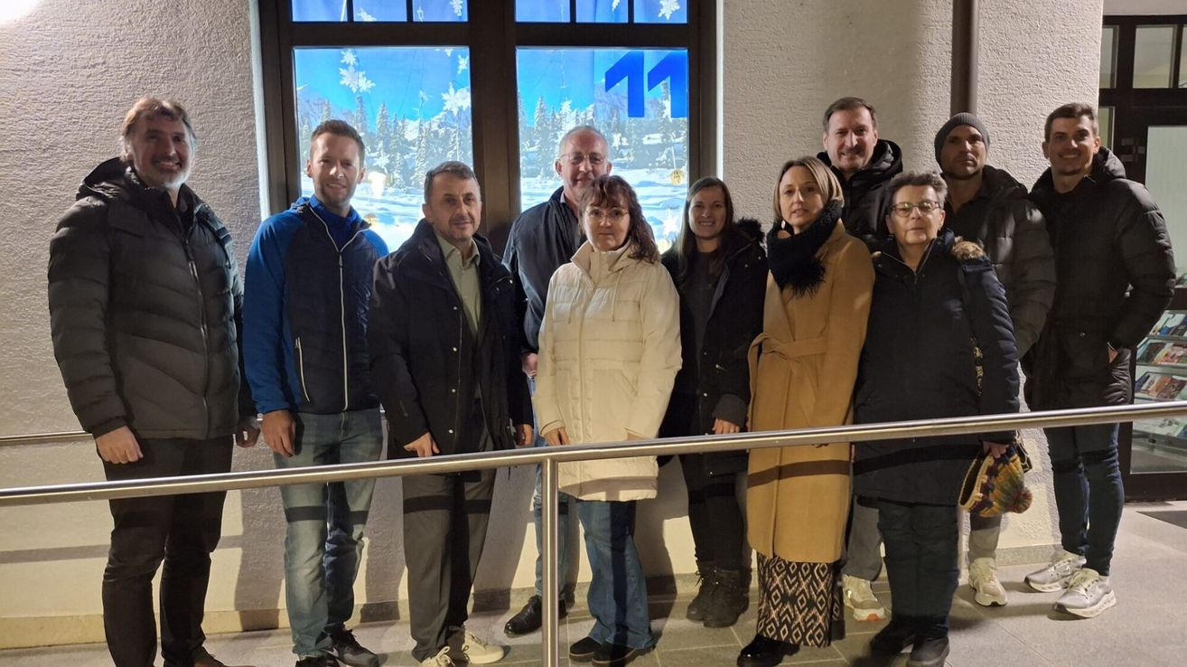 Sieben Menschen stehen zusammen vor einer Glasscheibe, möglicherweise für ein Foto. Sie alle lächeln. Die Glasscheibe hat die Nummer 11 und Schneeflocken darauf. Dahinter ist eine weiße Wand.