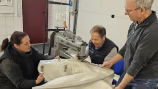 Drei Personen arbeiten in einem Raum mit einer Nähmaschine. Eine Person schaut auf einen großen weißen Stoff, während zwei andere zusehen.