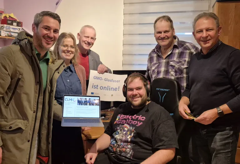 Eine Gruppe von Menschen lächelt für ein Foto. Ein Mann mit Headset sitzt in einem Stuhl mit Laptop. Ein Schild dahinter sagt GMO-Glasfaser ist online! Ein anderer Mann hält den Laptop.