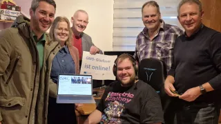 Eine Gruppe von Menschen lächelt für ein Foto. Ein Mann mit Headset sitzt in einem Stuhl mit Laptop. Ein Schild dahinter sagt GMO-Glasfaser ist online! Ein anderer Mann hält den Laptop.