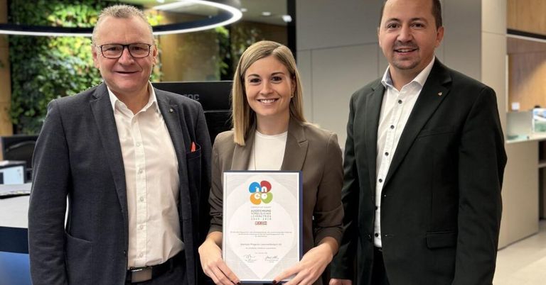 Eine Frau hält eine Auszeichnung in der Hand und steht zwischen zwei Männern, alle lächeln und posieren für ein Foto im Büro.