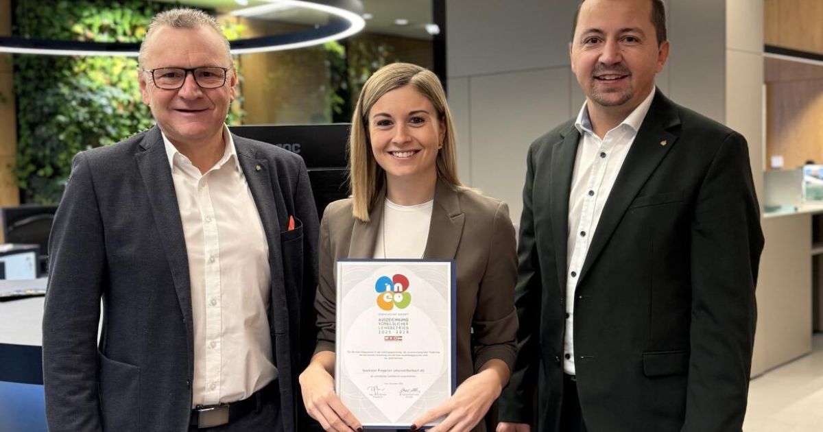 Eine Frau hält eine Auszeichnung in der Hand und steht zwischen zwei Männern, alle lächeln und posieren für ein Foto im Büro.