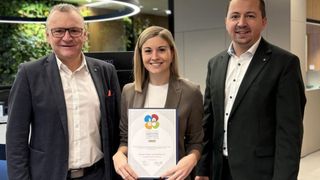 Eine Frau hält eine Auszeichnung in der Hand und steht zwischen zwei Männern, alle lächeln und posieren für ein Foto im Büro.