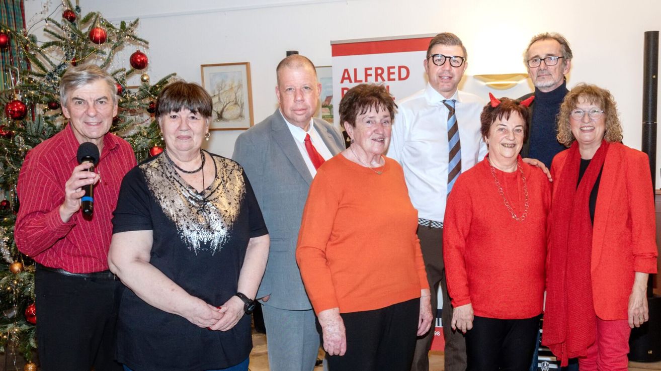 Fünf Personen stehen vor einem Weihnachtsbaum. Zwei Frauen und ein Mann tragen Halsketten. Hinter ihnen ist ein Plakat mit dem Text 'ALFRED'.