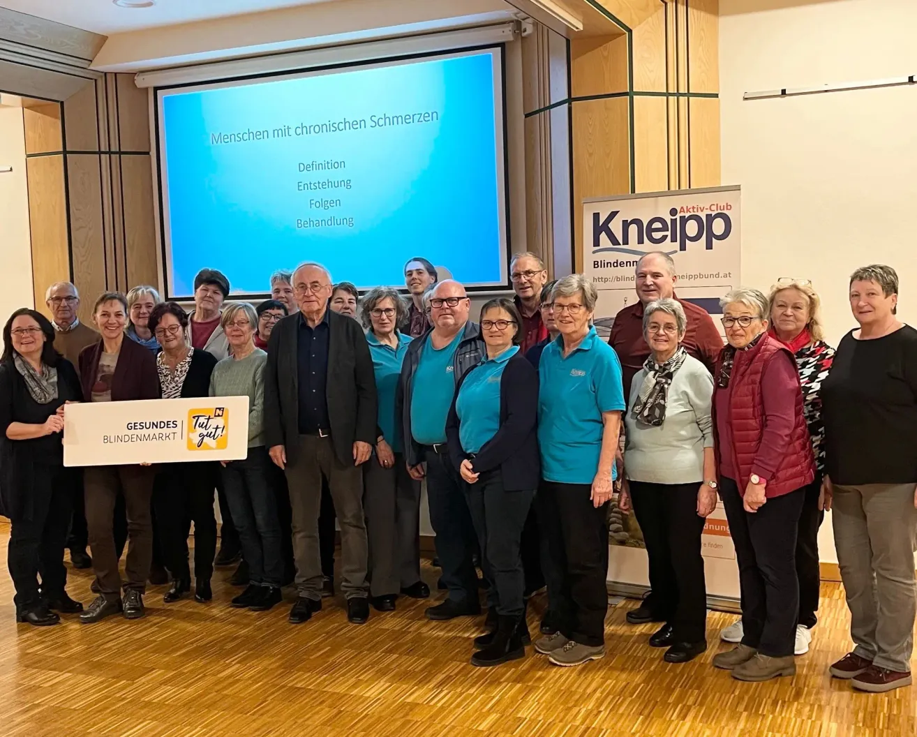 Gruppe von Menschen steht in einem Raum, einige tragen Brillen und Schuhe, ein Mann hält ein Banner, eine Beamerleinwand zeigt Text über chronische Schmerzen, ein Banner an der Wand und ein Holzboden.