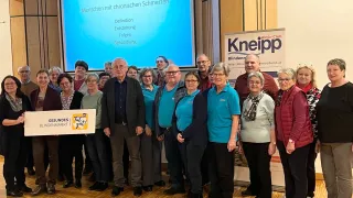 Gruppe von Menschen steht in einem Raum, einige tragen Brillen und Schuhe, ein Mann hält ein Banner, eine Beamerleinwand zeigt Text über chronische Schmerzen, ein Banner an der Wand und ein Holzboden.