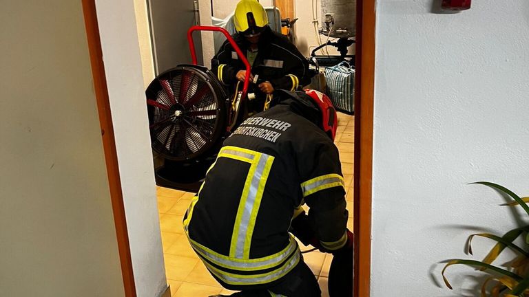 Zwei Feuerwehrleute arbeiten in einem Raum mit einem großen Ventilator. Ein Feuerwehrmann justiert ein Werkzeug und trägt einen Helm. Der andere bückt sich und trägt eine reflektierende Weste. Der Boden ist gefliest.