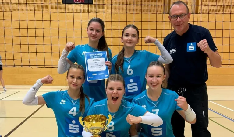 Eine Gruppe junger Frauen in blauen Trikots posiert für ein Foto mit einer Trophäe, eine hält ein Zertifikat. Ein Mann steht hinter ihnen.