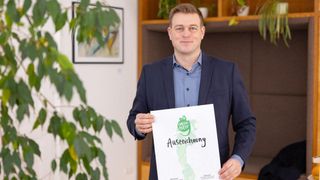 Ein Mann hält ein Zertifikat vor einer Pflanze und lächelt. Dahinter ist eine Wand mit einem Bilderrahmen.