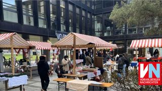 Ein Freiluftmarkt mit Ständen unter gestreiften Zelten, auf dem Menschen Artikel betrachten und kaufen, vor der Kulisse eines modernen Gebäudes.