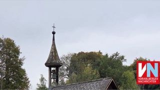 Ein Glockenturm mit einem Dachreiter und einem Kreuz darauf, der sich auf einem Gebäude mit einem Schieferdach befindet, umgeben von Bäumen.