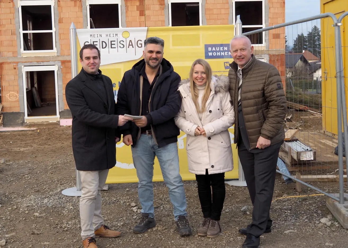 Vier Personen stehen vor einer Baustelle. Der Mann auf der linken Seite hält ein Stück Papier, und die Frau in der Mitte lächelt. Sie sind alle warm gekleidet für das kalte Wetter. Ein gelber Banner mit Text steht hinter ihnen.