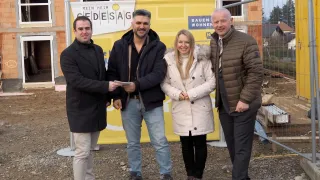 Vier Personen stehen vor einer Baustelle. Der Mann auf der linken Seite hält ein Stück Papier, und die Frau in der Mitte lächelt. Sie sind alle warm gekleidet für das kalte Wetter. Ein gelber Banner mit Text steht hinter ihnen.