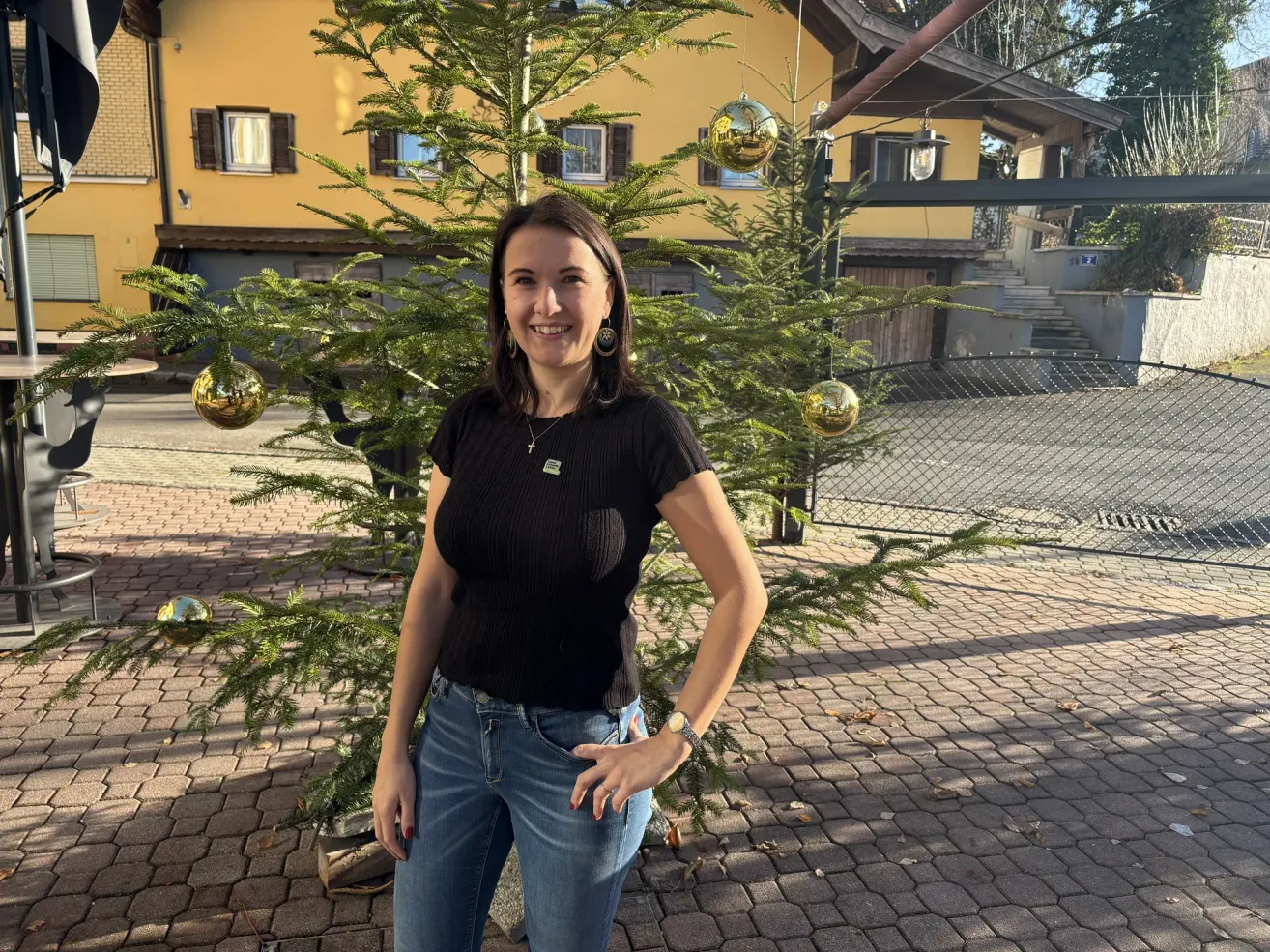 Eine Frau steht vor einem dekorativen Tannenbaum mit goldenen Ornamenten, lächelt und trägt ein schwarzes Oberteil und Jeans. Hinter ihr befindet sich ein gelbes Gebäude mit Fenstern und einer Treppe.