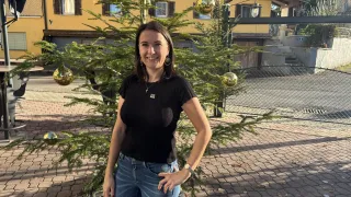 Eine Frau steht vor einem dekorativen Tannenbaum mit goldenen Ornamenten, lächelt und trägt ein schwarzes Oberteil und Jeans. Hinter ihr befindet sich ein gelbes Gebäude mit Fenstern und einer Treppe.