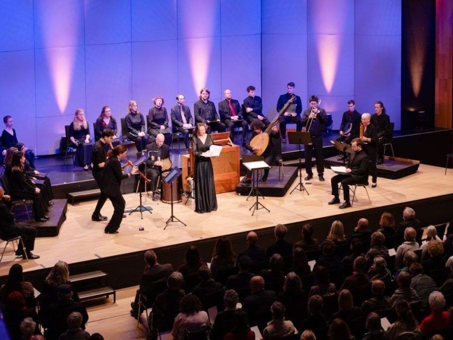 Ein klassisches Musikensemble spielt auf der Bühne mit Streich- und Blasinstrumenten. Eine Frau in einem schwarzen Kleid steht vor einem Pult. Das Publikum sitzt im Vordergrund.